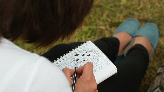 Young woman sitting on the grass outdoor and spending time on drawing pattern in her sketch book