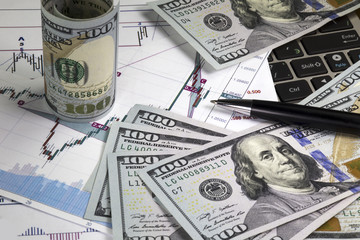 Graph with japanese candlesticks of euro dollar prices. Angled hundred us dollar bills above the chart. Rolled bill lying on graph diagonally. Notebook keyboard under the graph. Macro image.
