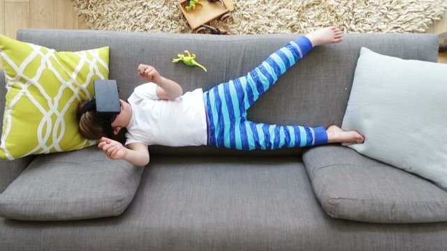 Aerial shot of a little boy lying on a sofa at home with his toy dinosaurs around him. He is using a virtual reality headset.