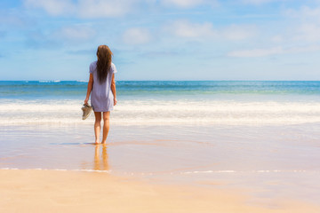 Young girl in dress and shoes in hand standing on beach and looking into sea.
