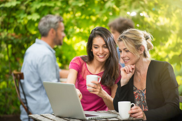 Two women friends sitting at a terrace cafe using a laptop
