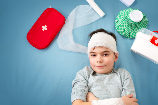 Child With Bandages Lying In Bed. Wounded Boy On Blue Background With First Aid Accessories.