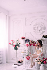 Beautiful young sexy woman , holding a glass and sitting in a chair, wearing white lingerie, room decorated with flowers. Perfect makeup. Beauty fashion. Eyelashes. Studio retouched shot.