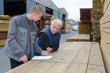 Men leaning on stack of wood studying clipboard - obrazy, fototapety, plakaty