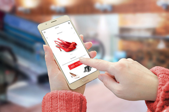 Shopping web site app on smart phone. Woman holding mobile device and buy red shoes. City shopping center in background.