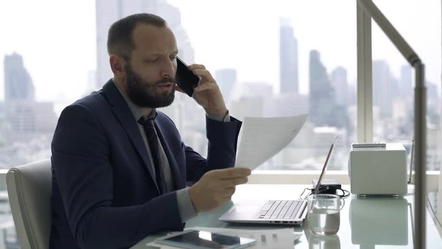 Young businessman with laptop and documents talking on cellphone in office 
