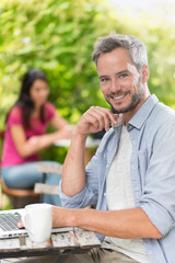 A gray haired man sitting at a terrace cafe and using a laptop