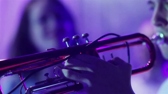 Man playing trumpet blue light neon close up. Performing wind instrument at stage in night club at new years party holding trumpet with finders and blowing - woman singing background shallow dof