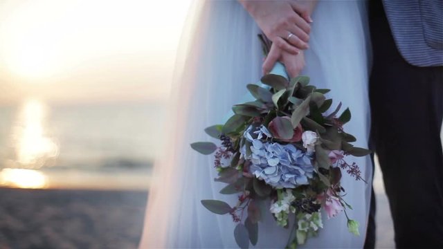Unusual wedding bouquet close up outdoors. Woman hands hold bridal bouquet made from flowers and green leaves at sunset sea view background. Floral design decoration. Newlyweds posing at beach no face