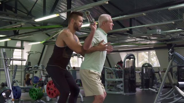 Side view of young male trainer controlling exercises of senior client at the gym. Caucasian bearded guy holding his hands under aged sportsman's arms. Old gray man training under supervision of