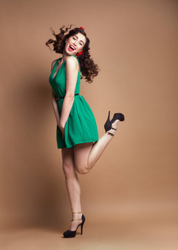 Curly Pin-up girl in green dress having fun and jumping on high heels at light brown rough background