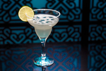 View of a Margarita Coctail in blue font, with a lemon slice on the edge of the glass. Colourful coctail on the black background