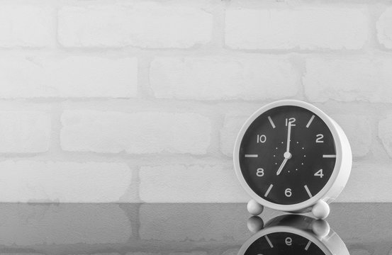 Closeup black and white alarm clock for decorate in 7 o'clock on black glass table and white brick wall textured background in black and white tone with copy space