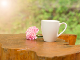 White cup of hot coffee with pink flower on wood table in natural green background in morning.