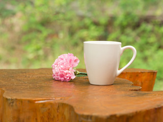 White cup of hot coffee with pink flower on wood table in natural green background in morning.