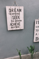 Vintage wooden board with motivational phrase hanging on the grey wall. Phrase - Dream the impossible, Seek the unknown, Achieve greatness.