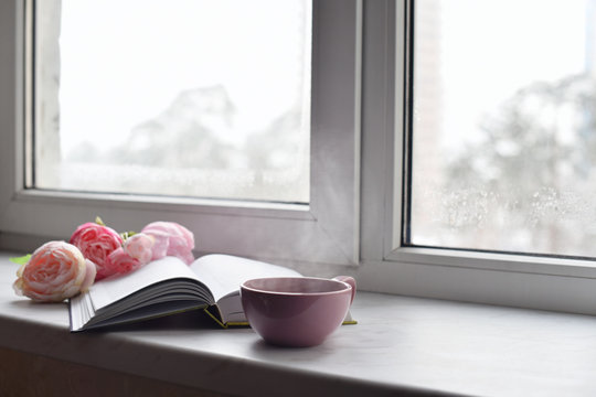 Cozy Home Still Life: Cup Of Hot Coffee, Spring Flowers And Opened Book With Warm Plaid On Windowsill Against Snow Landscape Outside. Springtime Concept, Free Copy Space