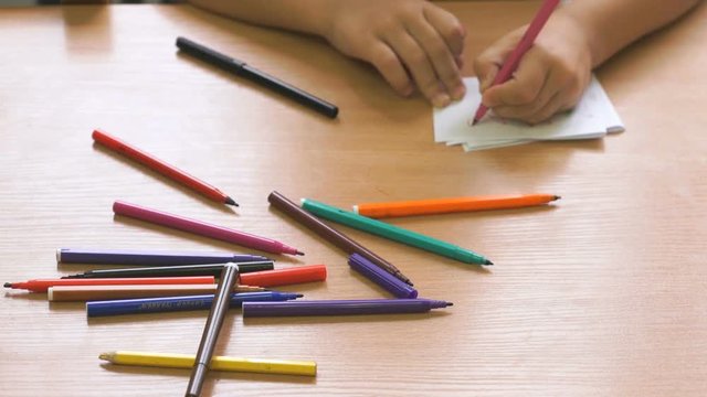 Little girl sitting at the desk draws the picture on the paper sheet using the multicolored felt pens. Close-up