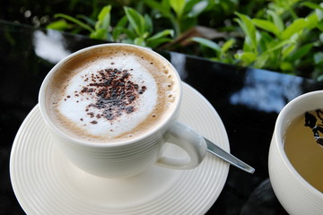 A white cup of cappuccino with spoon and chinese on glass table with green leaf background.