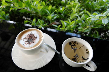 A white cup of cappuccino with spoon and chinese on glass table with green leaf background.
