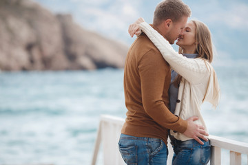 Young couple blond guy with short hair in a brown sweater and blue jeans and a blonde girl with straight long hair,spend time together,standing arm in arm on a white wooden pier near the sea in autumn