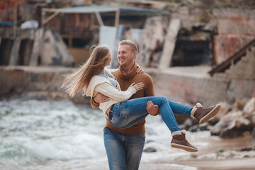 A couple in love,young woman ,long blond straight hair and a young man with short blonde hair in a brown sweater and blue jeans,the guy carries a girl on his hands,walking on the beach near the sea