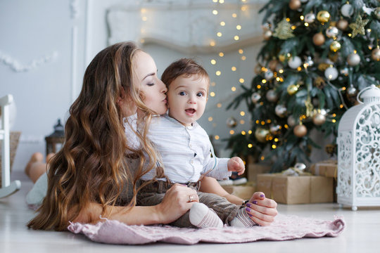Beautiful woman,dressed in a short white lace dress,holding New Year's holiday lie on the floor next to a white elegant Christmas tree with his young son,Christmas portrait of mother and baby