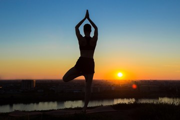 Silhouette of sporty woman practicing yoga in the park at sunset - tree pose, vrikshasana. Sunset light, golden hour. Freedom, health and yoga concept