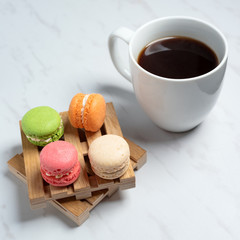 Sweet and colourful macaroons served on little wooden pallets with cup of coffee on a marble texture background. Traditional french dessert.