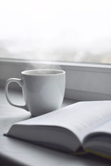 Cozy home still life: cup of hot coffee and opened book on windowsill against snow landscape outside. Winter holidays, Christmas time concept, free copy space