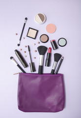 Aerial view of a purple leather make up bag, with cosmetic beauty products spilling out on to a pastel colored background