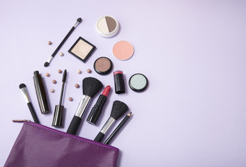 A purple leather make up bag, with cosmetic beauty products spilling out on to a pastel colored background, with blank space at side