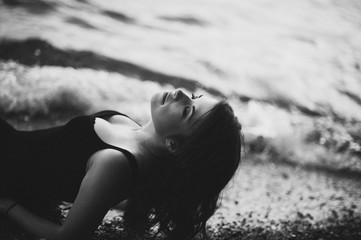 Beautiful girl wearing the black dress lying on the pebbles on the beach in the waves