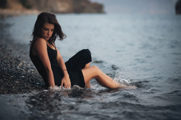 Beautiful girl wearing the black dress lying on the pebbles on the beach in the waves
