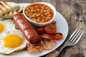 Traditional full English breakfast with fried eggs, sausages, beans, mushrooms, grilled tomatoes and bacon on wooden background
