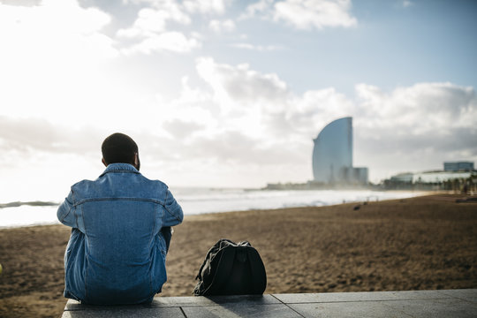 Spain, Barcelona, back view of young traveler sitting at the beach