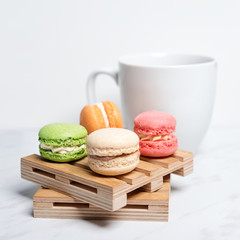 Sweet and colourful macaroons served on little wooden pallets with cup of coffee on a marble texture background. Traditional french dessert.