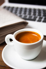 Cup of espresso with laptop and newspaper on wooden desk, closeup, vertical edit