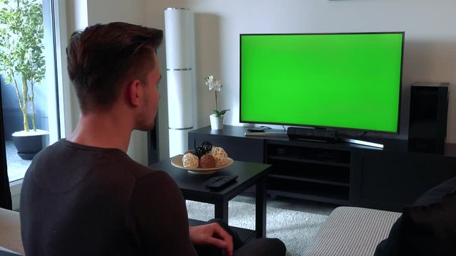 A young, handsome man watches a TV with a green screen, then turns to the camera, smiles and shows a thumb up