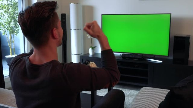 A man watches a TV with a green screen in a cozy living room and begins to celebrate (shout cheerfully, clap his hands and triumphantly raise his fists)