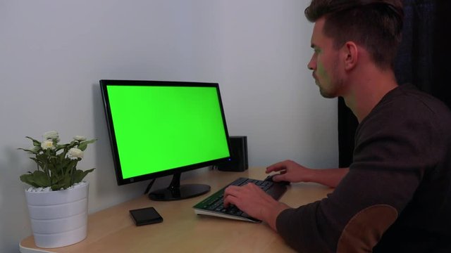 A young, handsome man types on a computer with a green screen