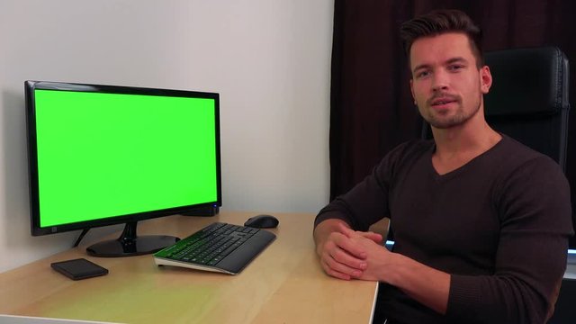 A young, handsome man sits at a desk in front of a green computer screen and explains something to the camera
