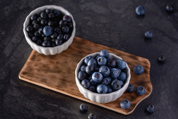 Bowls with blueberries on rustic wooden board on black background