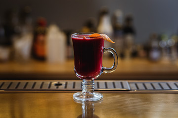 A transparent cup of raspberry mulled wine with orange peel on a bar stand. Selected focus, small depth of field, film grain effect. 