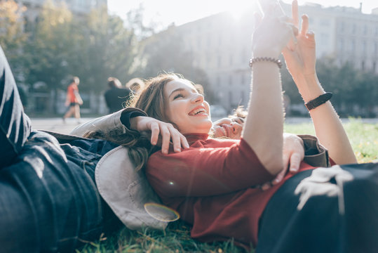 Young beautiful couple in love using smart phone together outdoor in the city lying on the grass in a park - interaction, love, technology concept