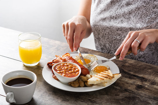 Woman eating a traditional English breakfast with fried eggs, sausages, beans, mushrooms, grilled tomatoes, bacon, coffee and orange juice, on wooden table

