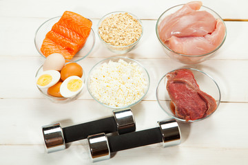 Protein diet, fish, cheese, eggs, meat, chicken and dumbbells on a white wooden background