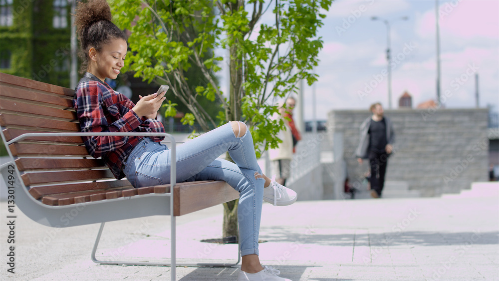 Wall mural pretty teenage girl using her mobile phone while sitting outdoors. casual style - jeans and checkere