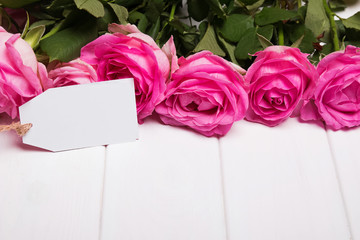 Beautiful pink roses and empty paper card on the white wooden ta