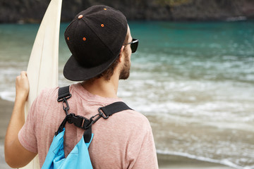 Stylish young bearded surfer wearing sunglasses and snapback looking at blue sea waves while standing on sandy beach with his bodyboard. Sports, extreme, hobby, active lifestyle and adventure